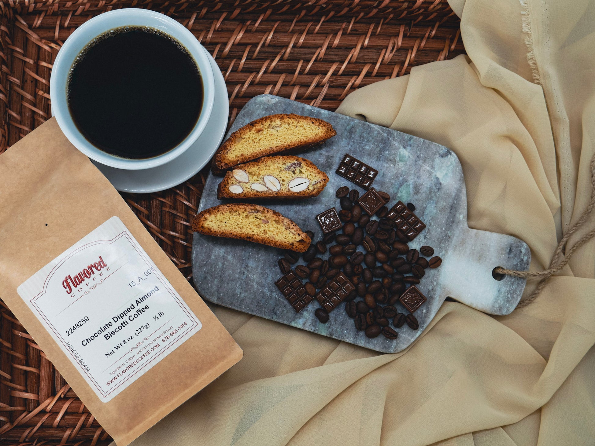 Cup of coffee, biscotti, and chocolate on a marble board with a package labeled 'Flavors' on a woven surface.