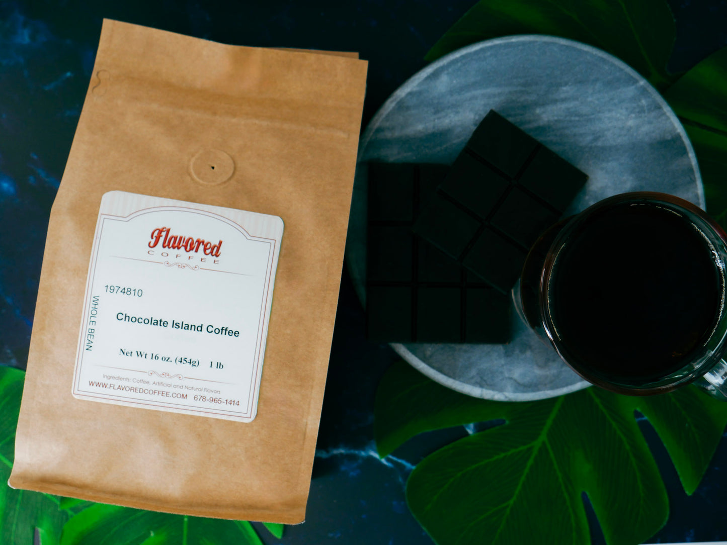 Bag of Chocolate Island coffee next to a mug with a dark cup of coffee, chocolate bars, on a grey coaster and dark background with green leaves.