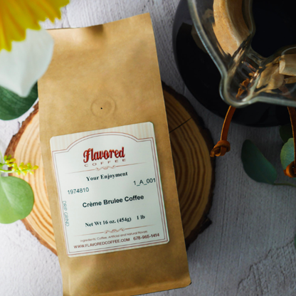 A bag of Flavored Coffee Crème Brulee Coffee in a brown kraft paper package with a white label, styled on a wooden slice coaster and a glass carafe nearby.