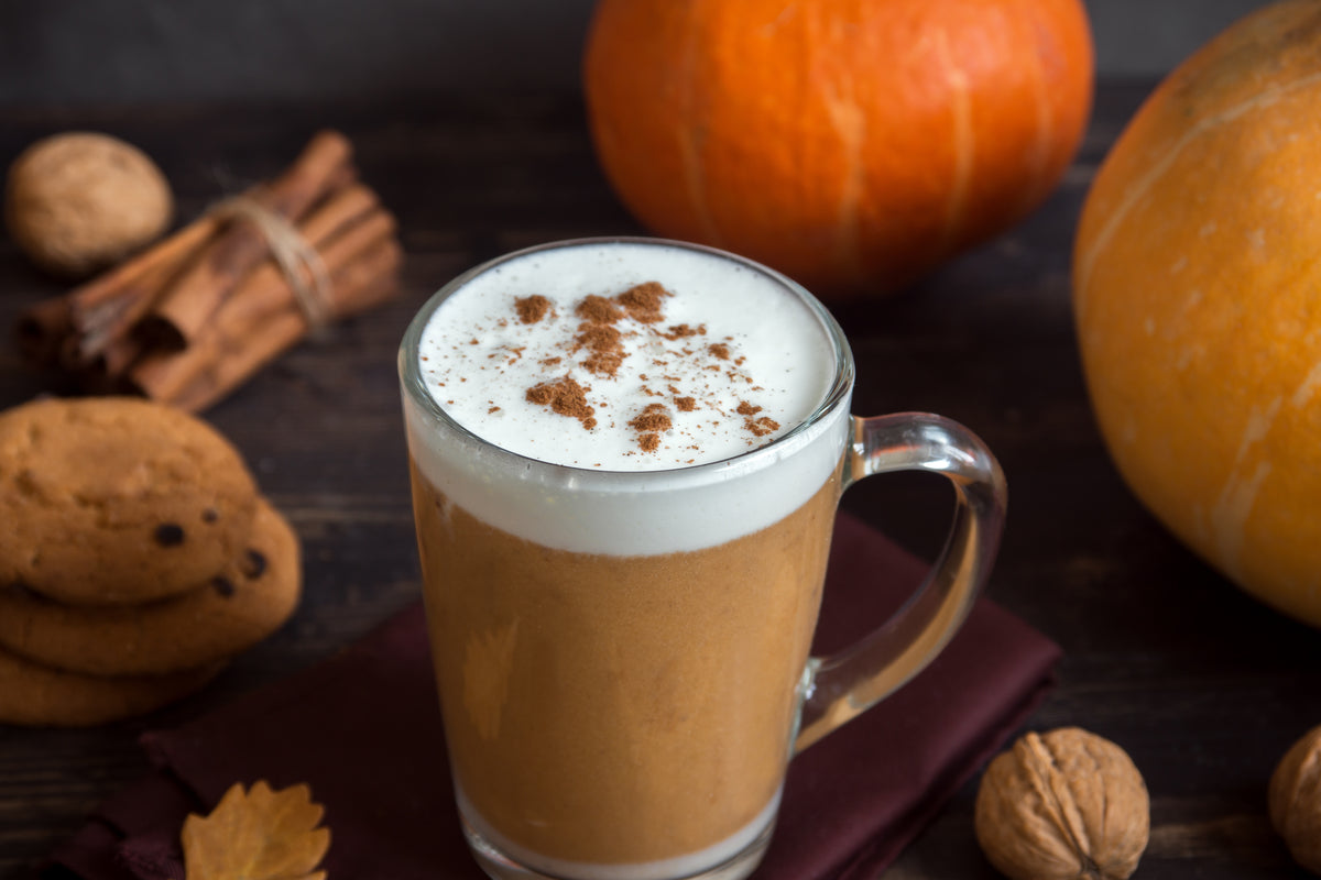 A glass mug filled with a warm pumpkin spice latte topped with whipped cream and cinnamon dusting, set on a dark wooden table surrounded by cookies, walnuts, cinnamon sticks, and pumpkins.