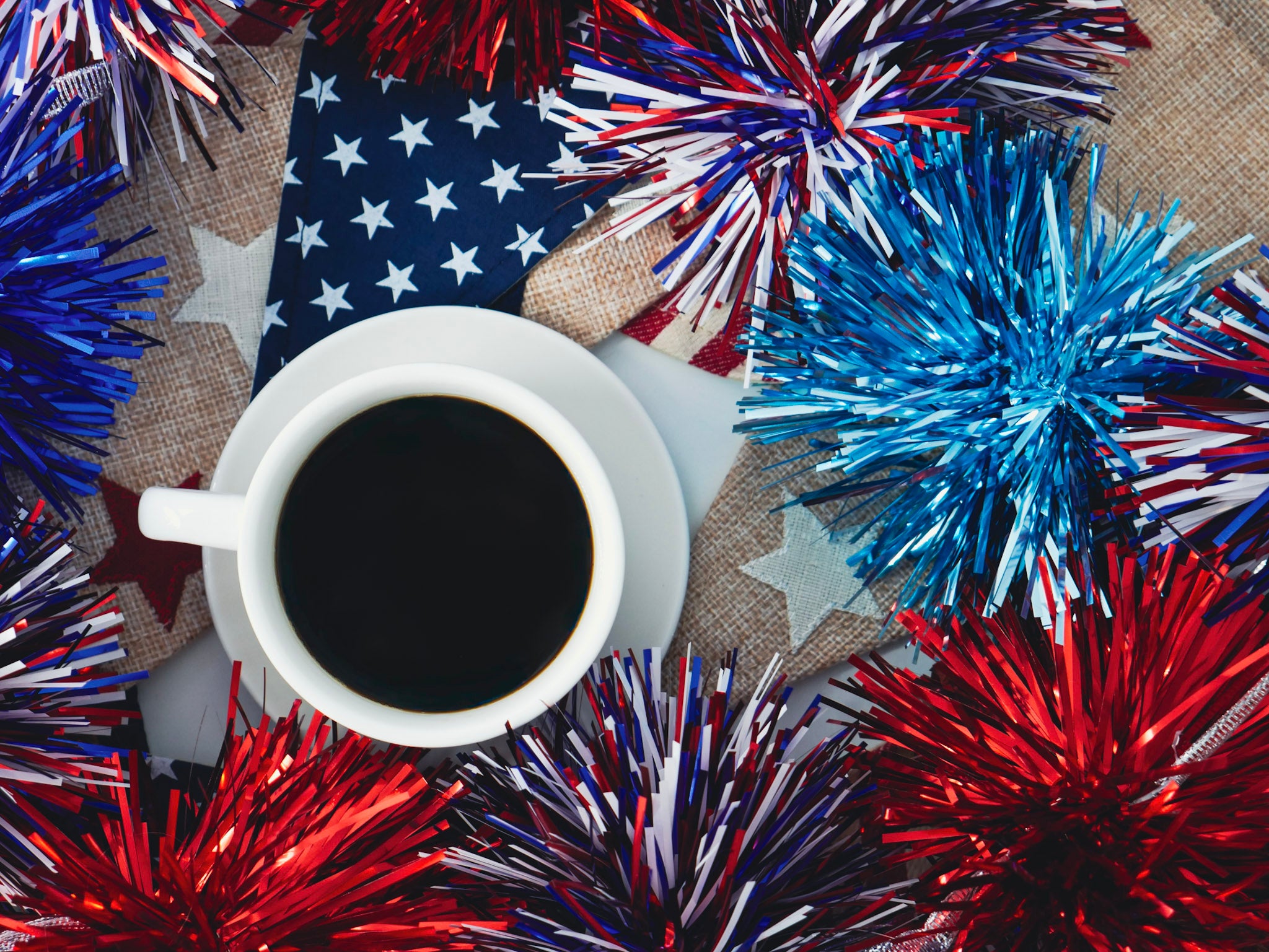 A white ceramic coffee cup filled with black coffee sits on a saucer, surrounded by patriotic red, white, and blue tinsel decorations, a small American flag, and burlap fabric.