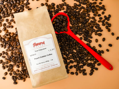 Bag of peach cobbler coffee on a peach colored background with a red coffee scoop and coffee beans