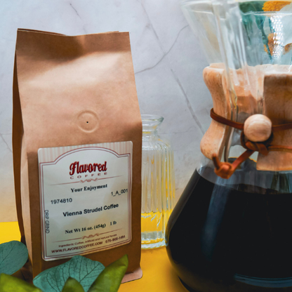 A brown Kraft bag of Flavored Coffee Vienna Strudel Coffee stands on a yellow surface next to a glass pour-over coffee maker with dark coffee, with green leaves in the foreground and a light marble-patterned background.