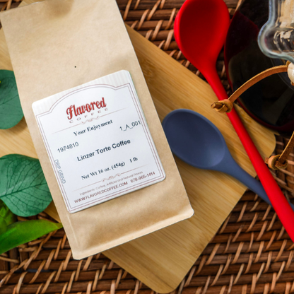 A 1 lb bag of Flavored Coffee Linzer Torte Coffee in a kraft paper pouch, styled on a wooden cutting board with a bamboo mat, a red spoon, a blue spoon, and green leaves.