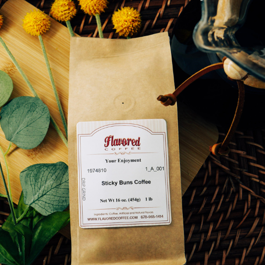 A kraft paper bag of Flavored Coffee Sticky Buns Coffee sits on a wooden surface with decorative yellow flowers, green leaves, with a glass pour over coffee next to the bag, and a wicker basket background.